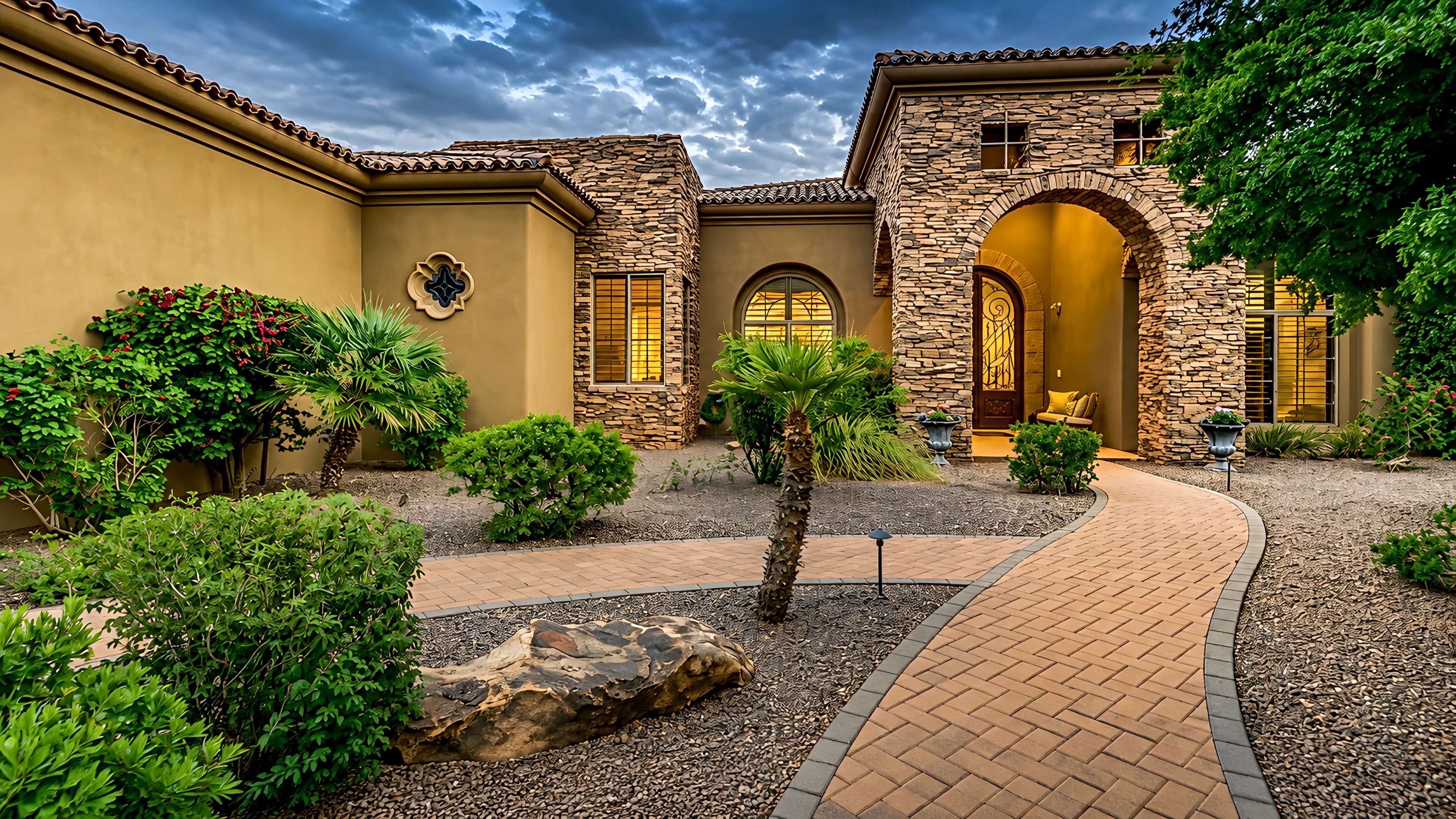 A paver walkway winding through palms and shaped shrubs to a lit stone entry